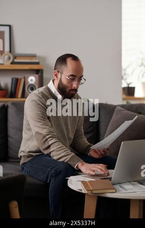 Young serious man in casualwear sitting on couch in living room and touching pad of laptop while checking online financial data Stock Photo