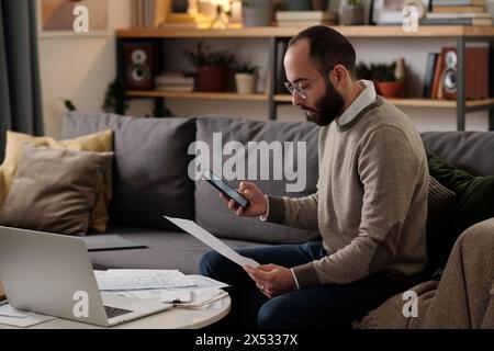 Bearded man in eyeglasses looking through sums in financial bill and using calculator in mobile phone while checking numbers Stock Photo