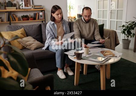 Young intercultural couple in casualwear sitting on couch in living room and looking through unpaid financial bills by table Stock Photo