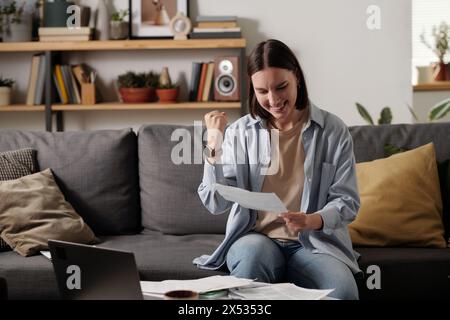 Happy young woman making yes gesture while holding clenched fist in front of herself and looking through paper document Stock Photo