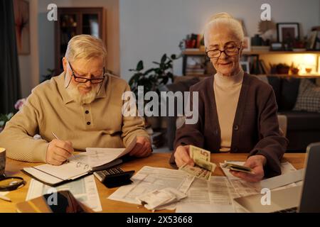 Aged man with financial bills in hand making notes on page of open notebook while sitting next to his wife counting dollar banknotes Stock Photo