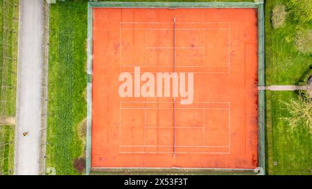 This aerial image features an unoccupied red clay tennis court, distinct with its bright playing surface and crisp white lines. Enclosed by lush green grass and a narrow path, the court stands out in a tranquil, natural setting, inviting a serene yet active vibe. Aerial View of an Empty Clay Tennis Court Surrounded by Greenery. High quality photo Stock Photo