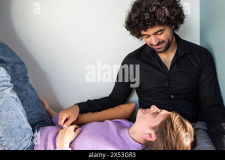 This warm photograph captures an intimate moment between a couple in the comfort of their home. A man with curly hair and a black shirt smiles fondly as he looks down at his partner, who lies relaxed in a lavender sweater, hand on his knee. The soft indoor lighting and their casual poses reflect a snapshot of everyday affection and companionship. Tender Moment: Couple Enjoying a Relaxed Connection at Home. High quality photo Stock Photo