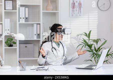 Excited Hispanic female doctor using virtual reality headset at her desk in a well-equipped medical office, exploring new technology in healthcare. Stock Photo