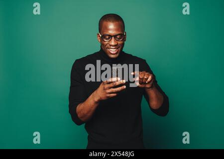 Stylish young man in casual clothing and glasses; standing against black background with green accent. Holding smartphone, happy and connected to the Stock Photo