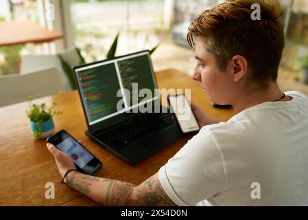 Person at wooden desk engages in intense multitasking with two smartphones displaying graphs, data analysis coding on laptop scene of modern Stock Photo