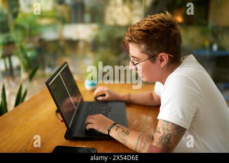 Transgender software developer programs at wooden desk. Focused on coding for mobile app, progressive workplace. Note tattoos, casual work attire Stock Photo