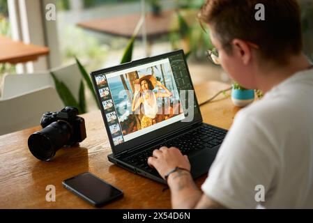 Photographer edits portrait on laptop in coffee shop, camera beside, focused work on digital editing, freelancer retouches image, using software tools Stock Photo