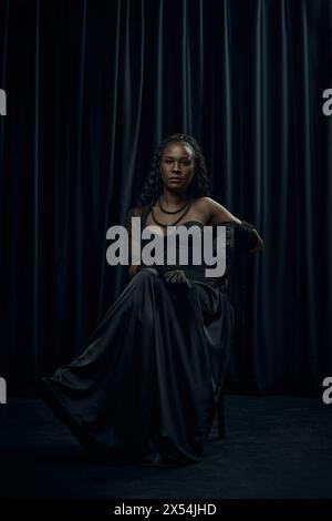African-American woman with long, braided hair sits confidently on chair, draped in old-fashion flowing black dress against dark curtain backdrop. Stock Photo
