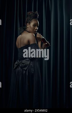 Young African-American woman, dressed as medieval person in classical black dress drinking soda against dark curtain backdrop. Stock Photo