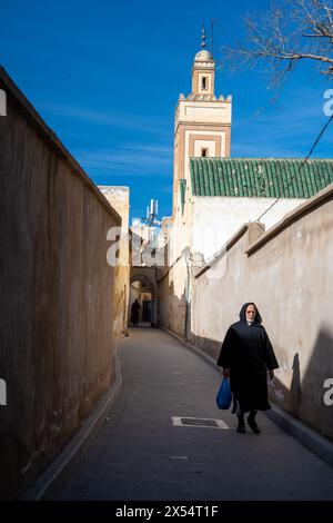 A man walks along a narrow street in Fez Medina with the minaret in the background on a clear day. Stock Photo