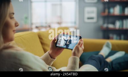 Beautiful Authentic Female is Using a Smartphone for Watching Online Video Lecture. She is Lying on a Couch Sofa at Home Living Room. University Student Studies From Home during Pandemic. Stock Photo