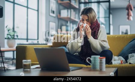 Ill Young Female Blowing Her Nose and Calling into Office with Video Phone Call from Cozy Living Room. Sick and Nauseous Woman in Warm Scarf is Using Laptop, Watching Movies. Stock Photo