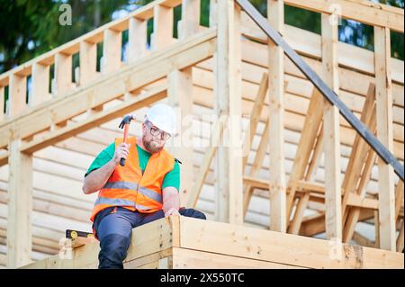 Carpenter constructing two-story wooden frame house. Bearded man in glasses hammering nails into structure while wearing protective helmet, construction vest. Concept of modern ecological construction Stock Photo