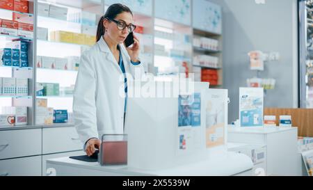 Pharmacy Drugstore Counter: Portrait of Helpful Caucasian Female Pharmacist Talking on the Mobile Phone with the Customer, Giving Medicine Advice, Uses Computer, Checks Prescription Availability Stock Photo