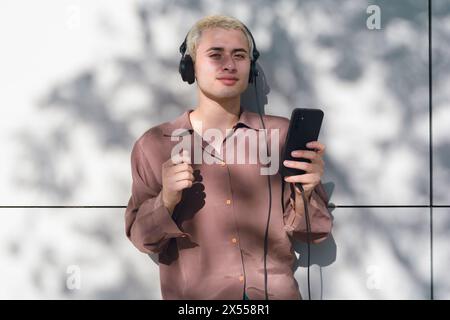 A young latin man with blonde hair is holding a cell phone and wearing headphones. He is standing in the shade and he is enjoying his music and lookin Stock Photo