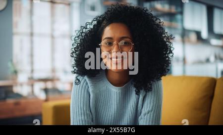 Portrait of a Beautiful Authentic Latina Female with Afro Hair Wearing Light Blue Jumper and Glasses. She Looks to the Camera and Smiling Charmingly. Successful Woman Resting in Bright Living Room. Stock Photo