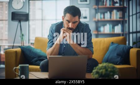 Handsome Caucasian Man Working on Laptop Computer while Sitting on a Sofa Couch in Stylish Cozy Living Room. Freelancer Working From Home. Browsing Internet, Using Social Networks, Being Serious. Stock Photo