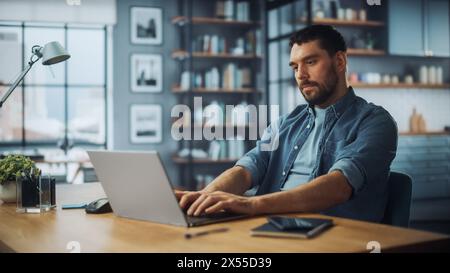 Handsome Caucasian Man Working on Laptop Computer while Sitting on a Sofa Couch in Stylish Cozy Living Room. Freelancer Working From Home. Browsing Internet, Using Social Networks, Being Serious. Stock Photo