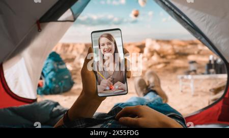 POV of a Tourist is Lying in a Tent and Using Smartphone for Making a Video Call to a Happy Smiling Girlfriend or Colleague. Traveller Resting in Tent on Top of a Mountain and Flying Hot Air Balloons. Stock Photo