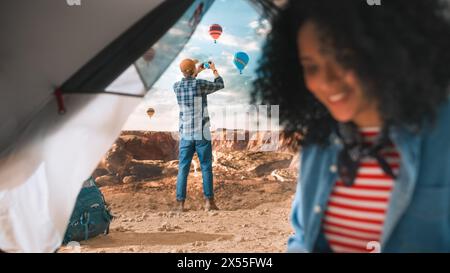 Diverse Adventurous Couple Mountain Hikers Living in Nature in Great Outdoors. Multiethnic Female Traveller with Afro Hairstyle Using Laptop Computer in a Tent on Top of Rocky Canyon. Stock Photo