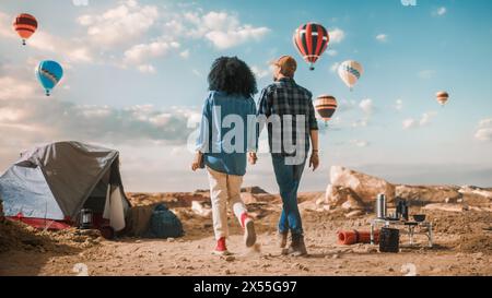 Young Diverse Tourist Couple Hiking and Living in a Tent in Great Outdoors in Rocky Canyon Valley. Male and Female Backpacker Hold Hands and Look at Hot Air Balloon Festival in Mountain National Park. Stock Photo