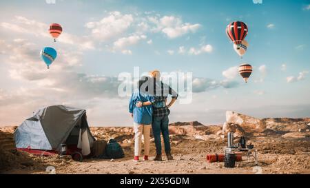 Young Diverse Tourist Couple Hiking and Living in a Tent in Great Outdoors in Rocky Canyon Valley. Male and Female Backpacker Hug and Look at Hot Air Balloon Festival in Mountain National Park. Stock Photo