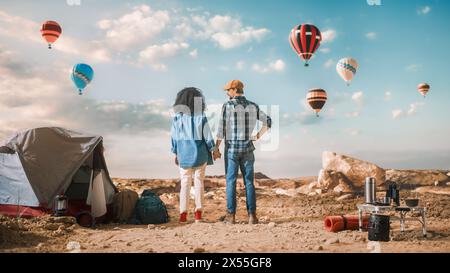 Young Diverse Tourist Couple Hiking and Living in a Tent in Great Outdoors in Rocky Canyon Valley. Male and Female Backpacker Hold Hands and Look at Hot Air Balloon Festival in Mountain National Park. Stock Photo
