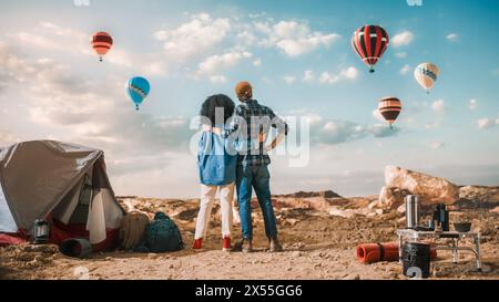 Young Diverse Tourist Couple Hiking and Living in a Tent in Great Outdoors in Rocky Canyon Valley. Male and Female Backpacker Hug and Look at Hot Air Balloon Festival in Mountain National Park. Stock Photo