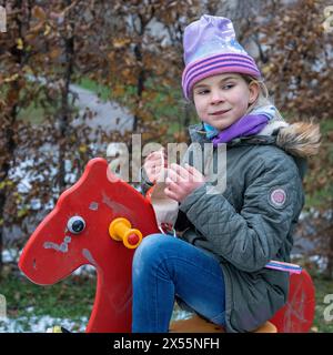 Girls love horses. When there is no animal around, children like to play in a playground where there is a red rocking horse. Healthy in cold weather. Stock Photo