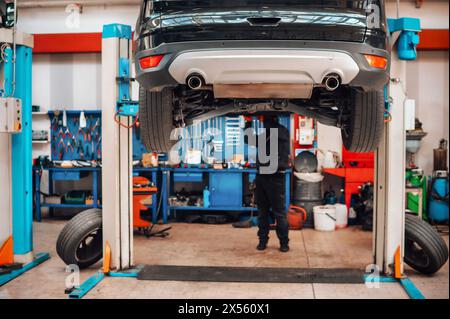 Anonymous male mechanic in uniform and gloves while standing working on lifted car front bottom in service workshop under lights Stock Photo
