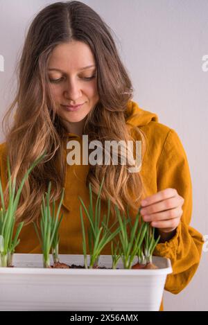 Woman, dressed in warm yellow hoodie, attentively tends to her thriving green onion garden, cultivated in white container at home. Stock Photo