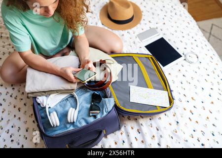A woman is sitting on the bed and packing her things in a travel bag, using a smartphone and looking for useful travel information. Stock Photo