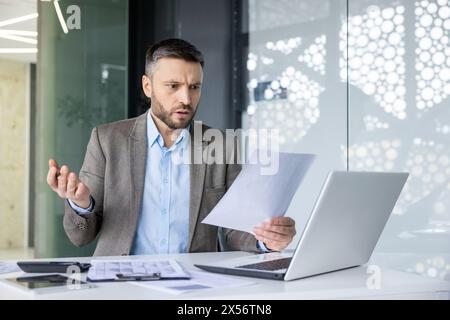 An adult male businessman looking confused while examining documents in a contemporary office setting. Features modern design and technology. Stock Photo