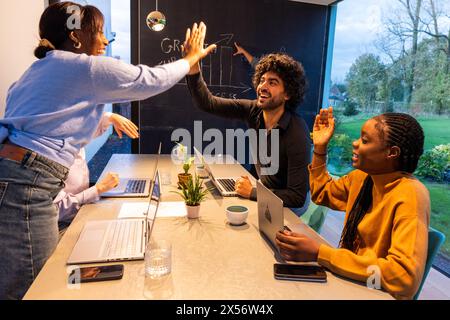 An exuberant moment in a modern office featuring a diverse team of three: an Asian woman, a Middle Eastern man, and a Black woman. They are gathered around a sleek, marble table filled with laptops and coffee mugs, celebrating a triumph. The Asian woman stands, giving a high-five to the seated Middle Eastern man, while the Black woman cheers them on. A chalkboard with a positive growth chart adds to the scene's upbeat and professional atmosphere. Diverse Team Celebrating Success in Modern Office. High quality photo Stock Photo