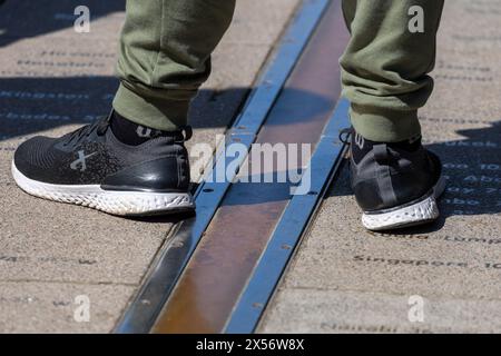 London, England - July 19, 2022: Pair of feet with black sneakers standing on both sides of meridian line on ground at Greenwich observatory. Stock Photo