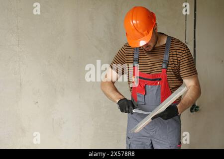 Worker in overalls and safety hard hat holds and prepares two spatulas for plastering concrete wall with putty using. Renovation apartment and construction new building concept. Copy place. Stock Photo