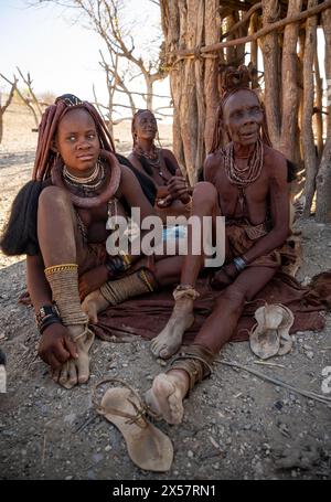 Young and old Himba woman, traditionally dressed, sit in front of a hut in the shade, near Opuwo, Kaokoveld, Kunene, Namibia Stock Photo