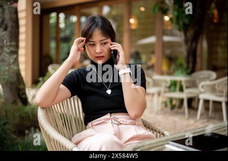 A beautiful serious Asian woman sits at an outdoors table of a cafe having a serious conversation on the phone with someone. people and wireless techn Stock Photo