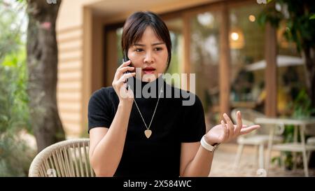 A beautiful serious Asian woman sits at an outdoors table of a cafe having a serious conversation on the phone with someone. people and wireless techn Stock Photo