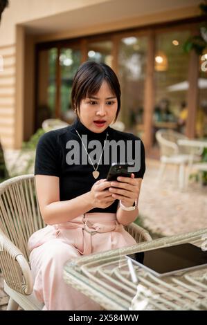 A shocked Asian woman looking at her smartphone screen with a stunning face, shocked with a message on her phone, sitting at an outdoor table of a caf Stock Photo