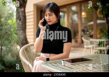 A beautiful serious Asian woman sits at an outdoors table of a cafe having a serious conversation on the phone with someone. people and wireless techn Stock Photo