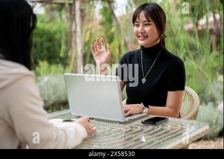 A cheerful young Asian woman in casual clothes talking with her friend while working remotely in the garden. discussion, interview, meeting Stock Photo