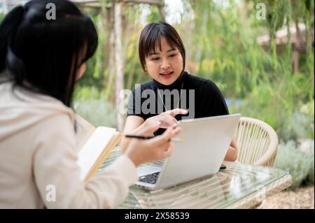 An attractive Asian woman in casual clothes working remotely in a garden with her colleague, discussing work and sharing ideas. Stock Photo
