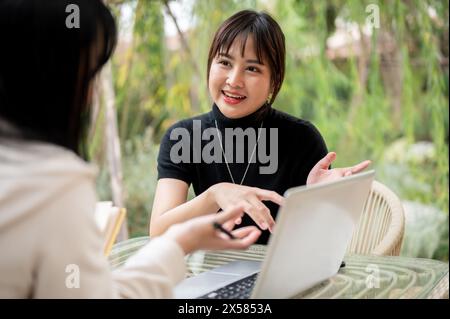 An attractive Asian woman in casual clothes working remotely in a garden with her colleague, discussing work and sharing ideas. Stock Photo