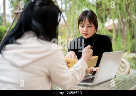 An attractive Asian woman in casual clothes working remotely in a garden with her colleague, discussing work and sharing ideas. Stock Photo