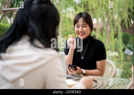 A cheerful young Asian woman in casual clothes talking with her friend while working remotely in the garden. discussion, interview, meeting Stock Photo