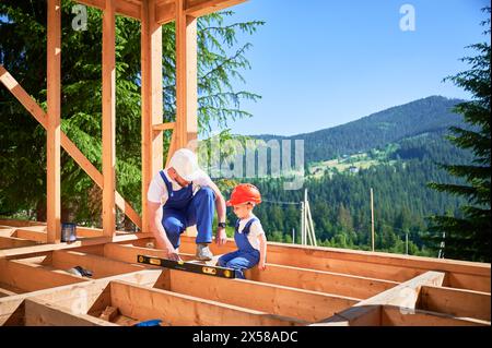 Father with toddler son building wooden frame house. Male builder demonstrating to his son how to measure level using spirit level on construction site on sunny day. Carpentry and family concept. Stock Photo