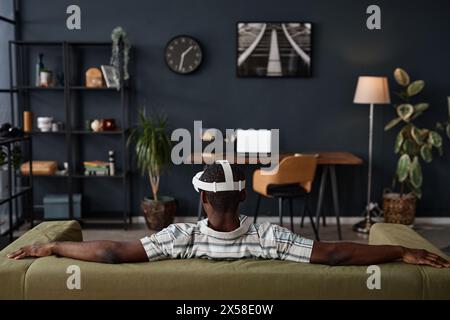 Rear view of unrecognizable Black man wearing VR headset sitting relaxed on couch in living room, copy space Stock Photo