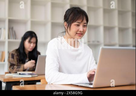 A positive, charming young Asian female university student is using her laptop computer while listening to a teacher lecture in the classroom. Stock Photo